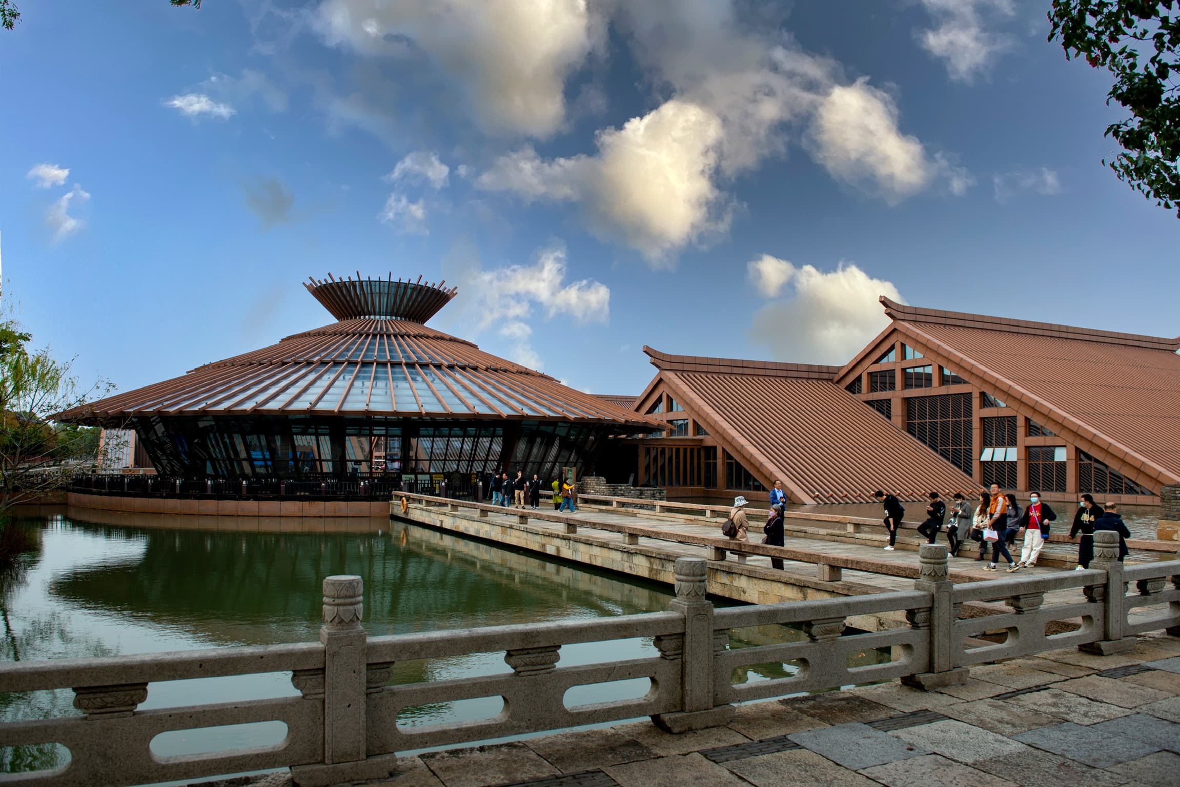 Modern Chinese building with a funnel-shaped roof and stone bridge over a reflective pond.