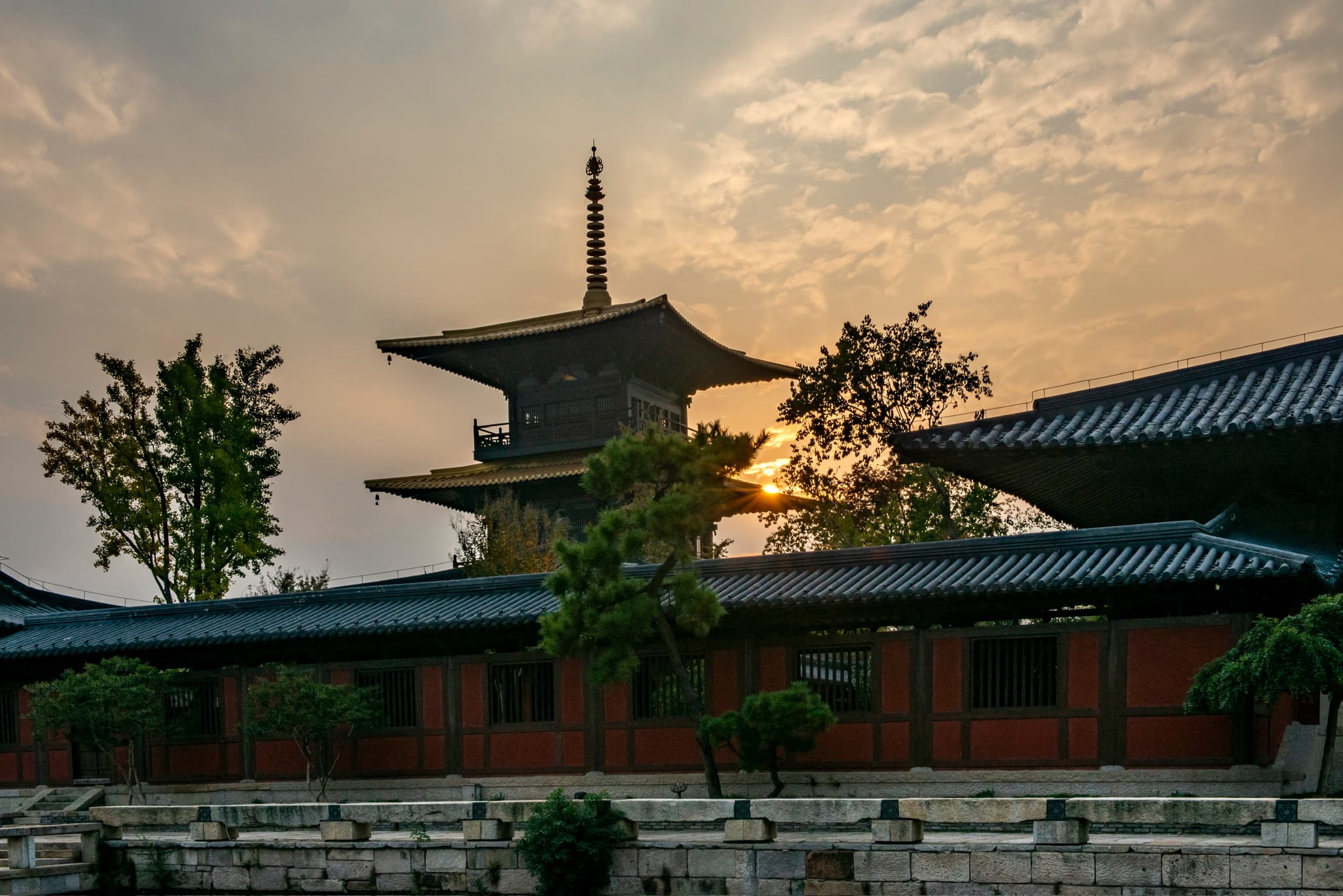 Traditional pagoda and tiled temple wall silhouetted against a golden, cloudy sunset sky.