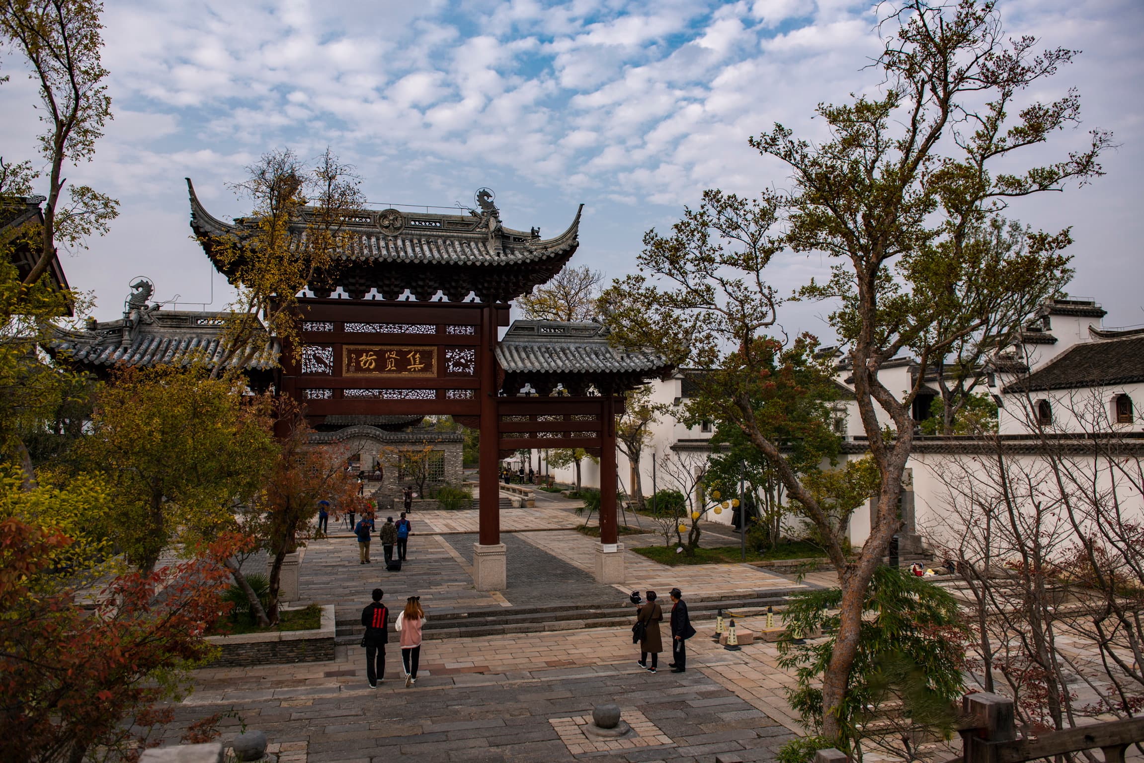 Traditional Chinese wooden gate in a stone courtyard with visitors and autumn trees.